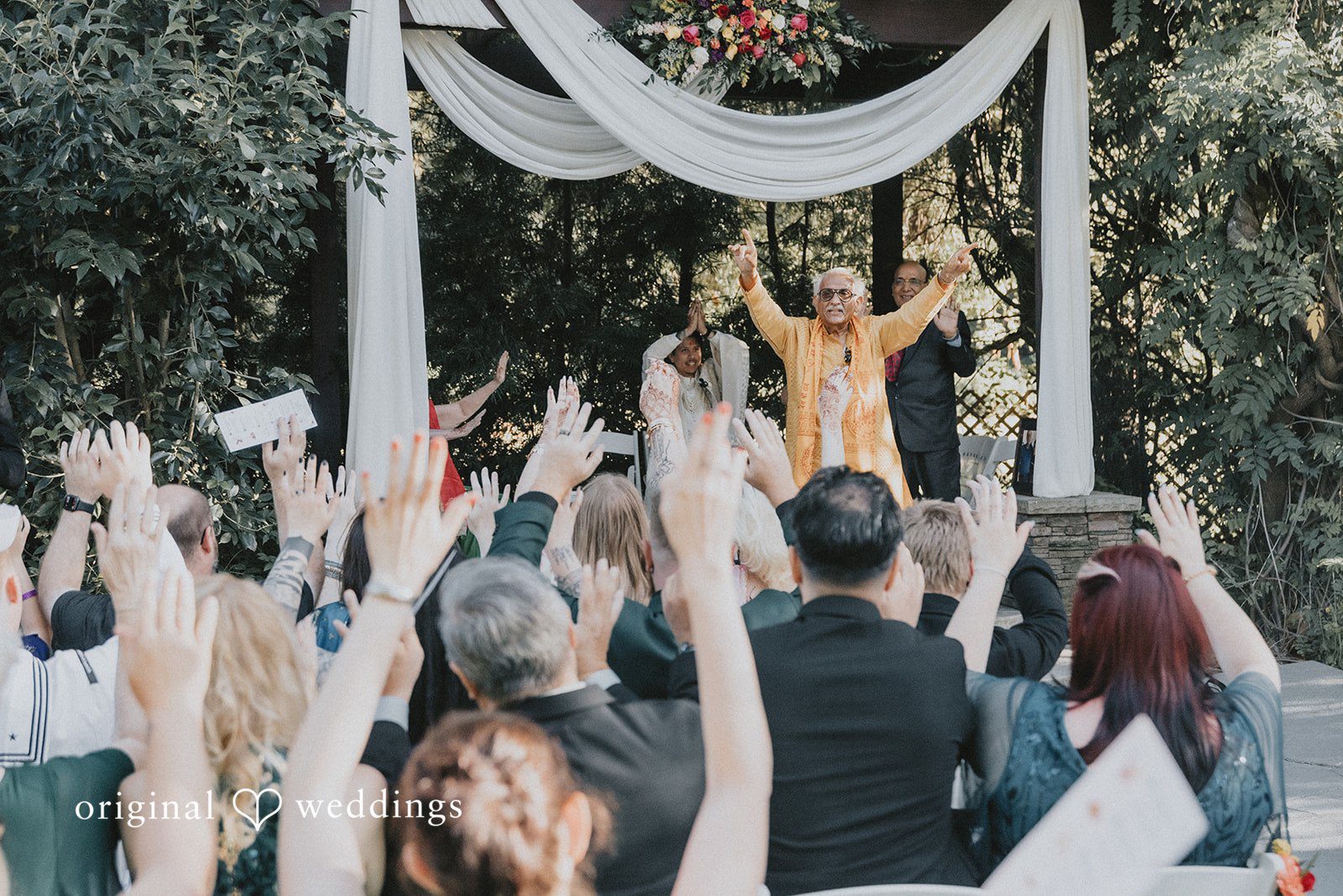 Wedding guests raising their hands during a ceremony under white drapery.