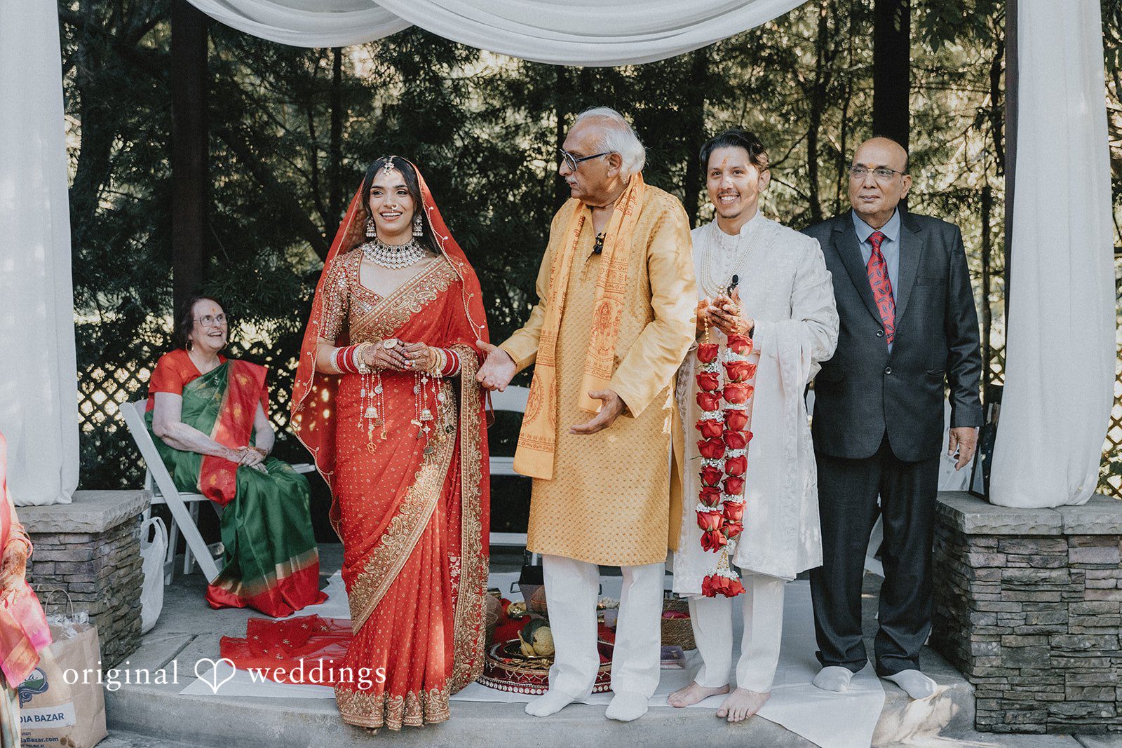 A bride in a red lehenga stands with three men in formal attire under a draped wedding altar.