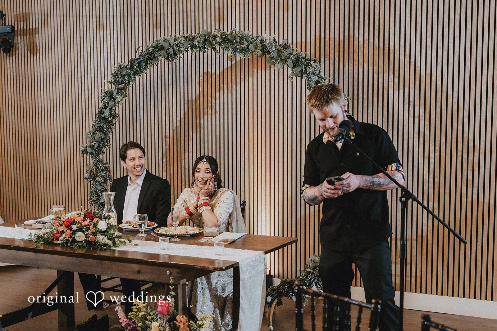 A speaker standing at a microphone next to a decorated head table at a wedding reception.