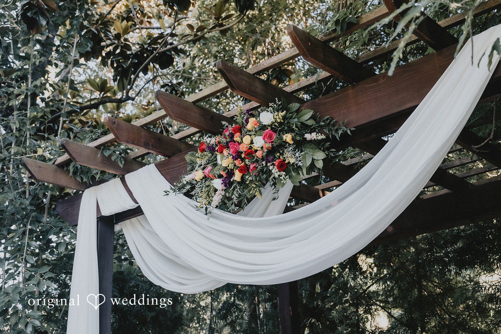 A dark wooden pergola structure draped with flowing white fabric and adorned with a colorful bouquet of fresh flowers.