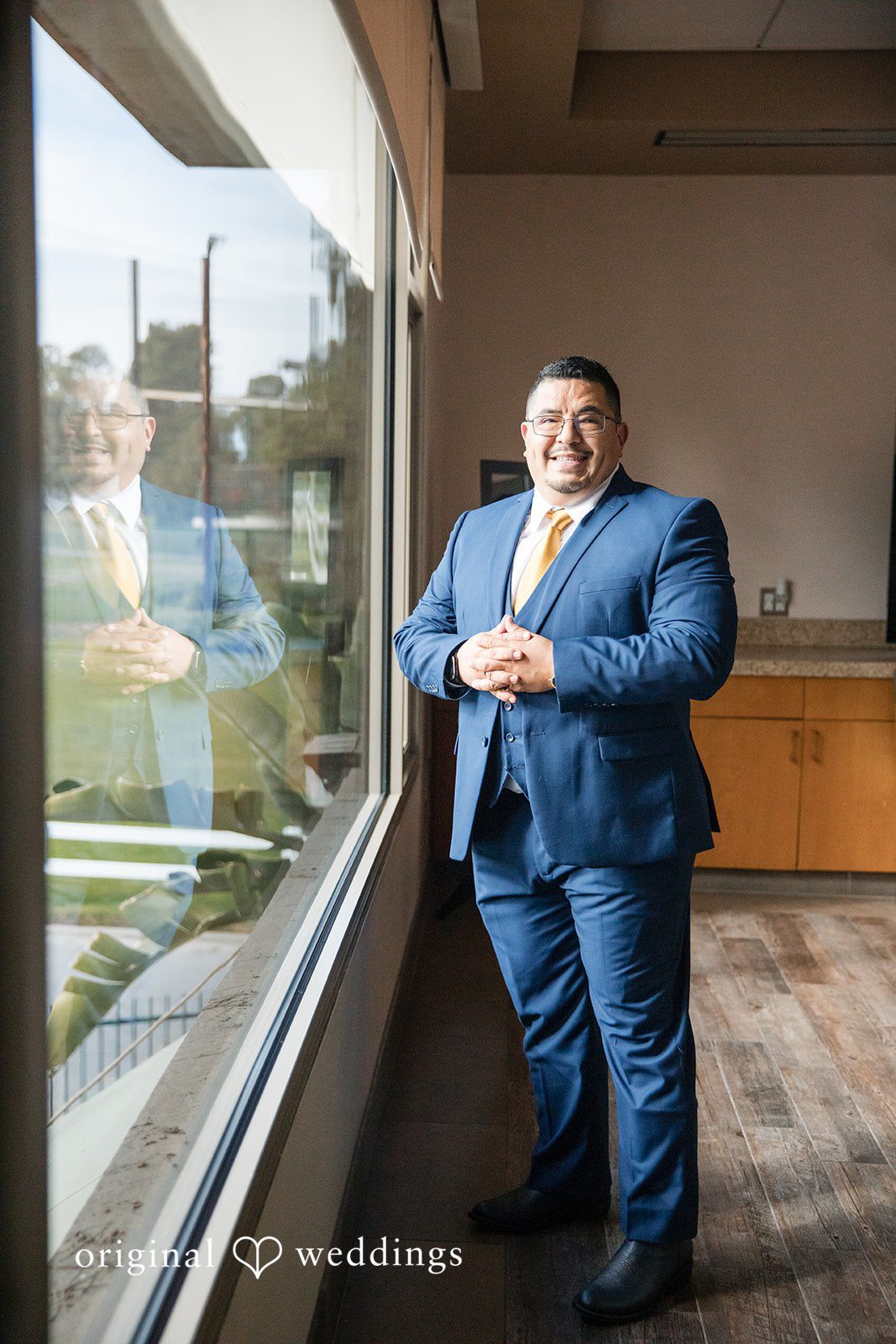 A groom in a blue suit standing near a window looking out.