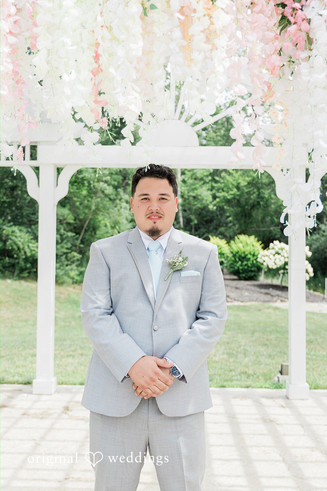 Alexander + Annalisse A groom in a light grey suit poses for a portrait against a backdrop of green trees.