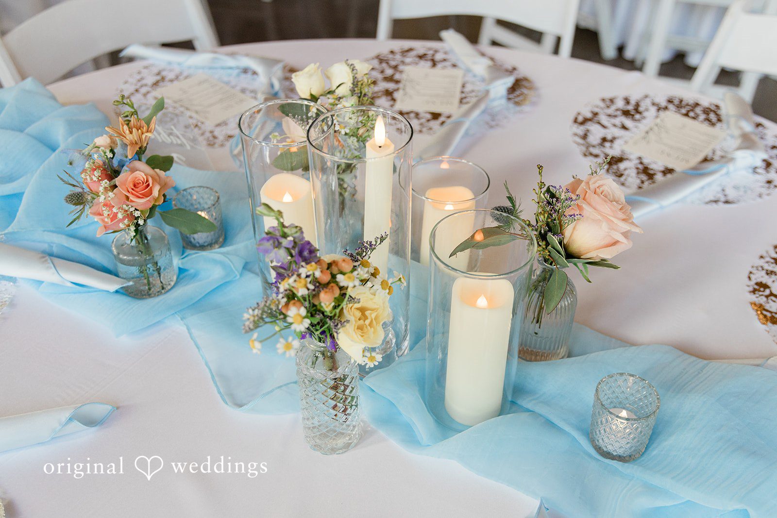 Alexander + Annalisse A close-up view of a reception table setting adorned with a light blue runner and glowing candles.