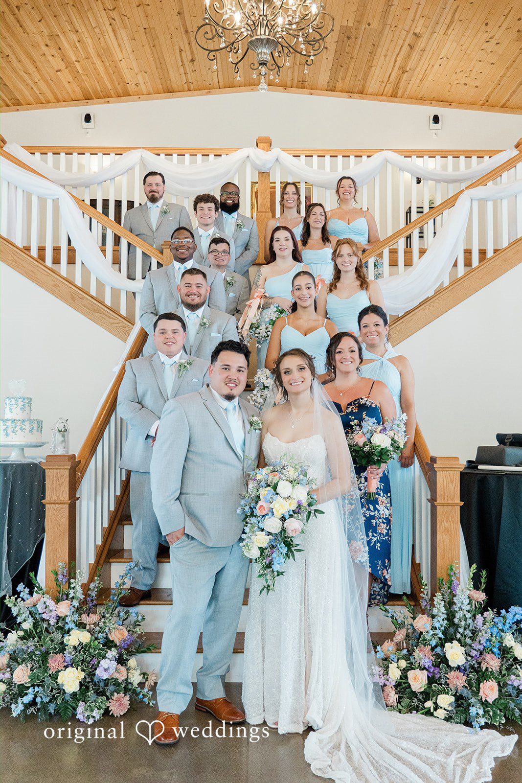 Alexander + Annalisse A large wedding party poses together on a grand wooden staircase inside a venue.