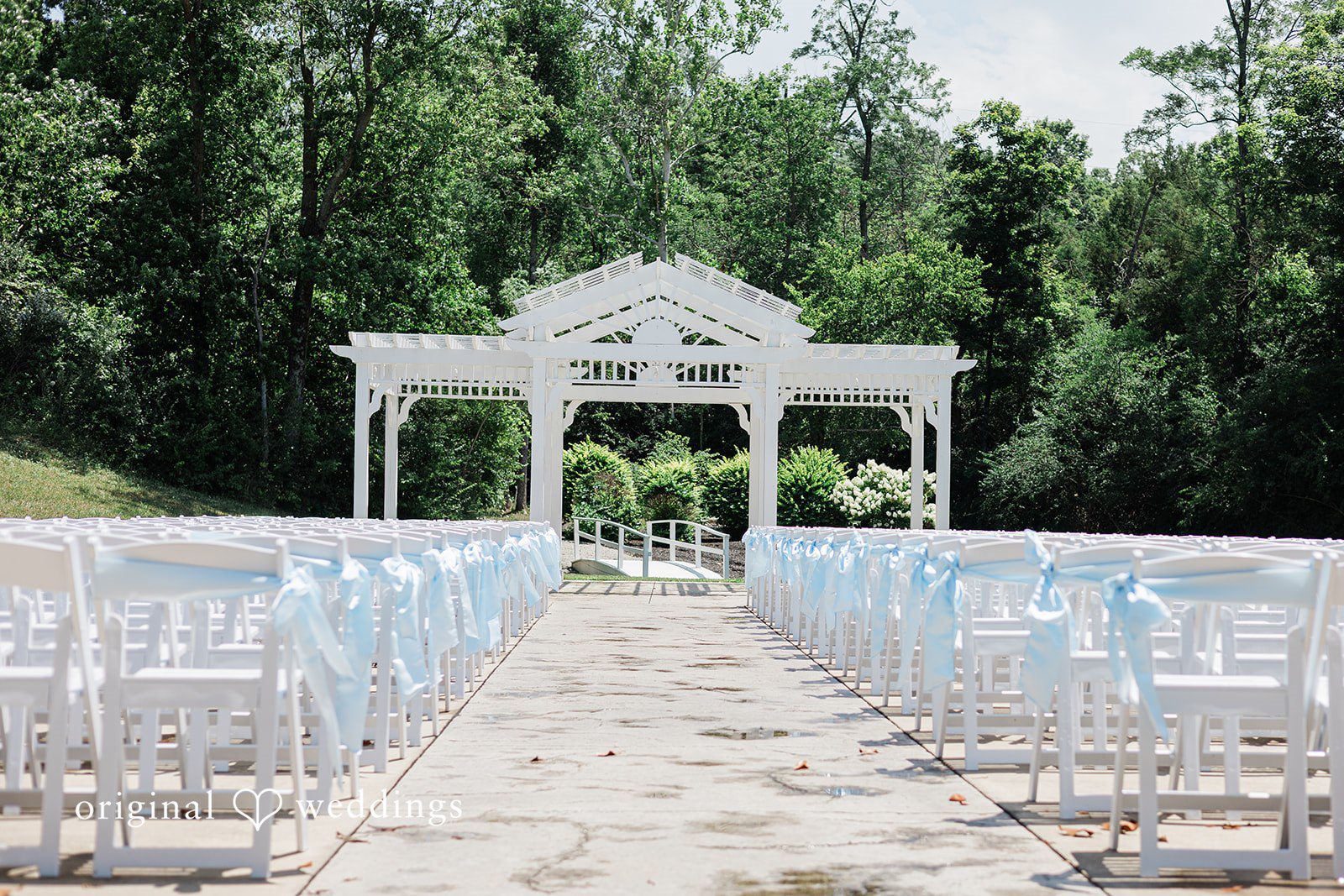 Alexander + Annalisse An elegant white wedding arbor stands at the end of an open aisle lined with rows of transparent ceremony chairs.