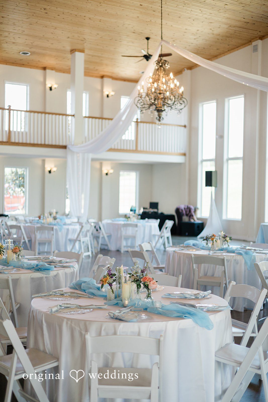 Alexander + Annalisse A bright reception hall is decorated with round tables, white chairs, and hanging chandeliers.