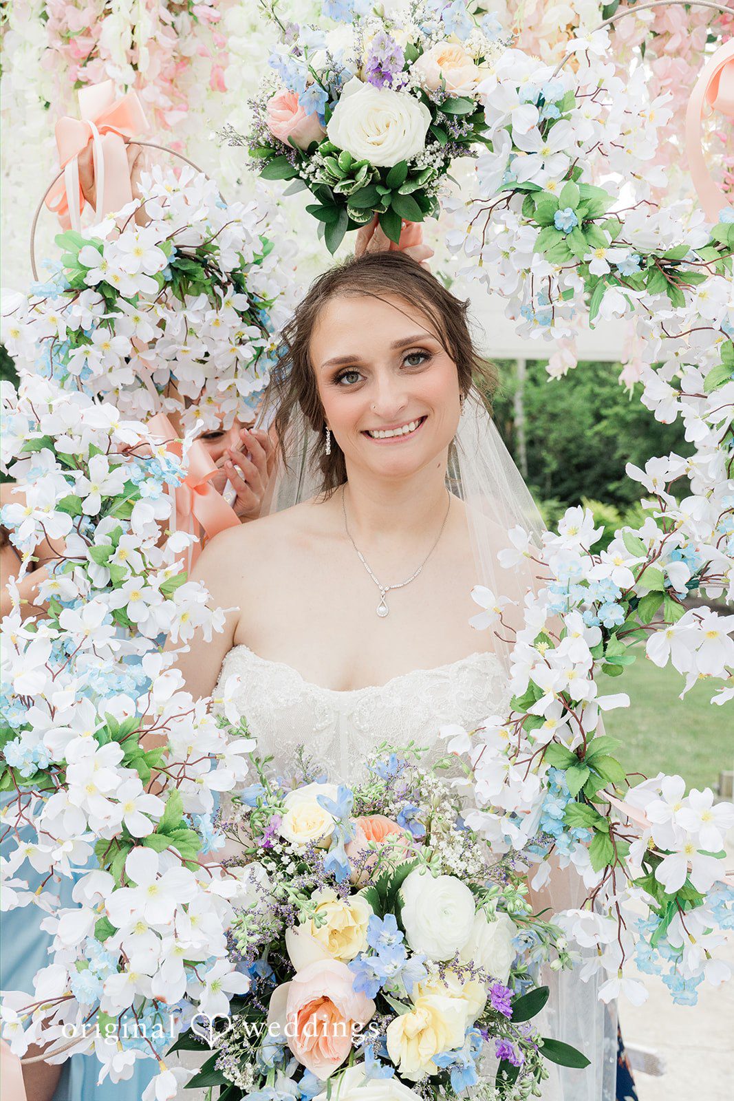 Alexander + Annalisse A bride smiles brightly while framed by a lush arrangement of white and blue flowers.