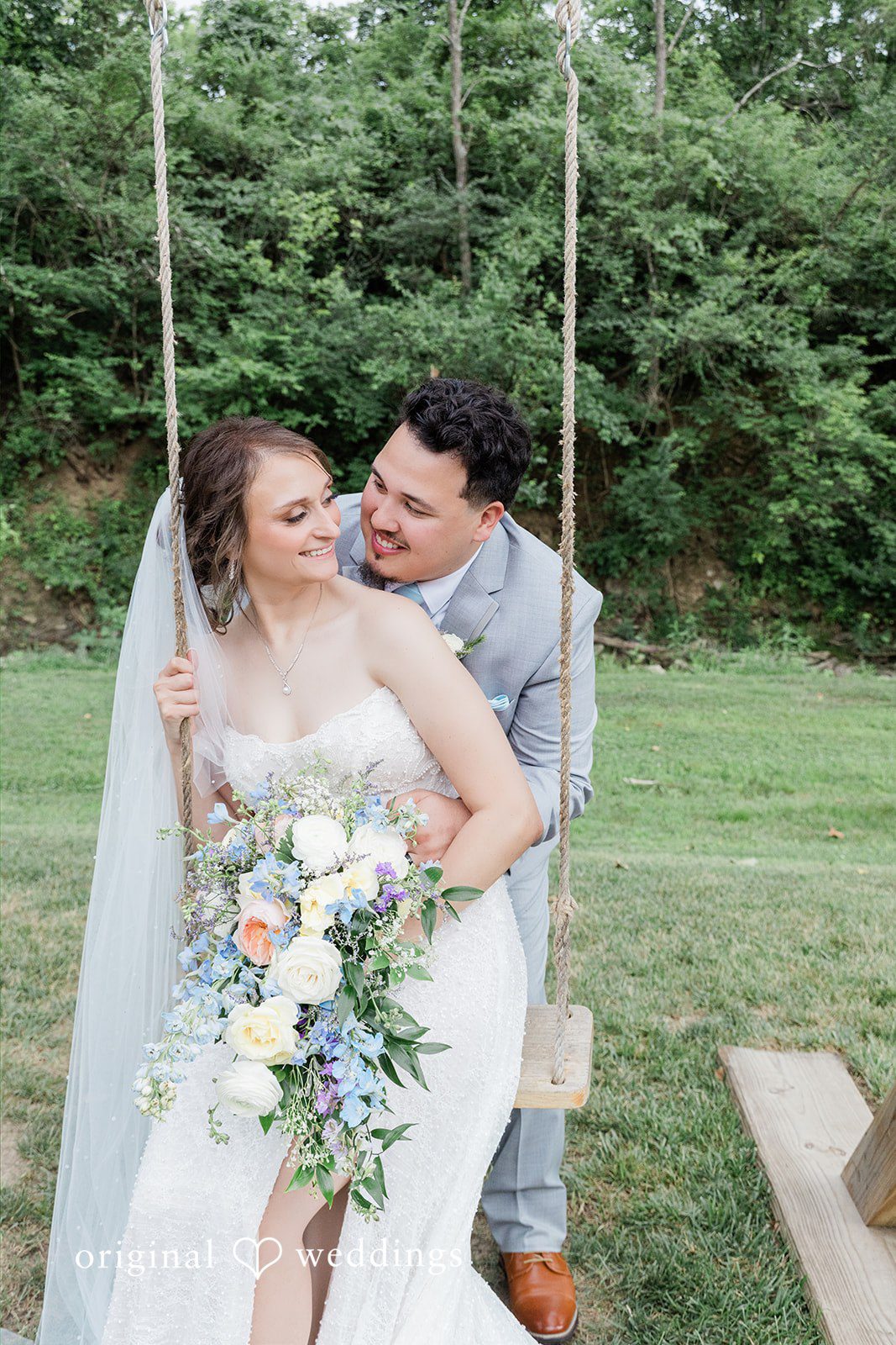 Alexander + Annalisse A bride holding a floral bouquet sits on a wooden swing while her groom stands beside her.