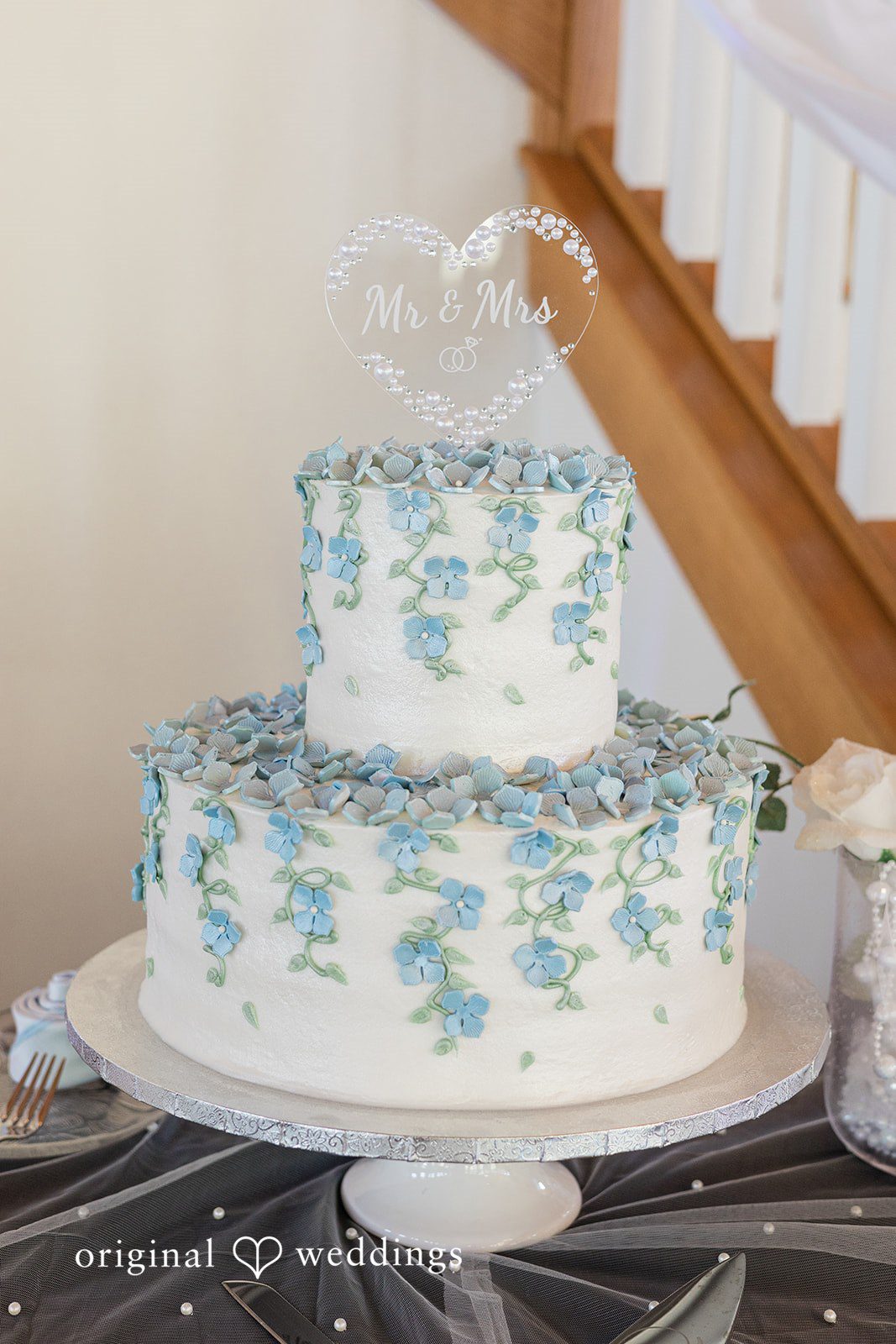 Alexander + Annalisse A three-tiered white wedding cake decorated with delicate blue and green floral patterns sits on a stand.