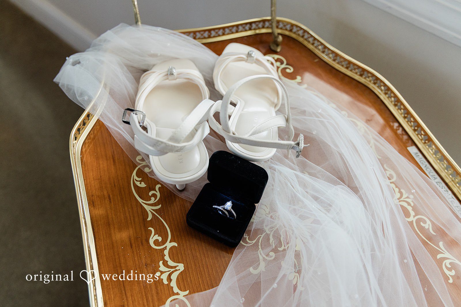 Alexander + Annalisse A bridal flat-lay features elegant white heels, jewelry, and a soft veil arranged on a wooden surface.