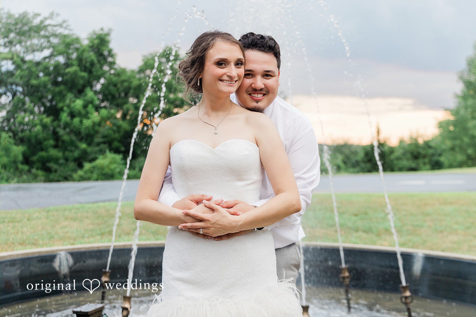 Alexander + Annalisse A bride and groom stand together near a fountain structure at dusk.