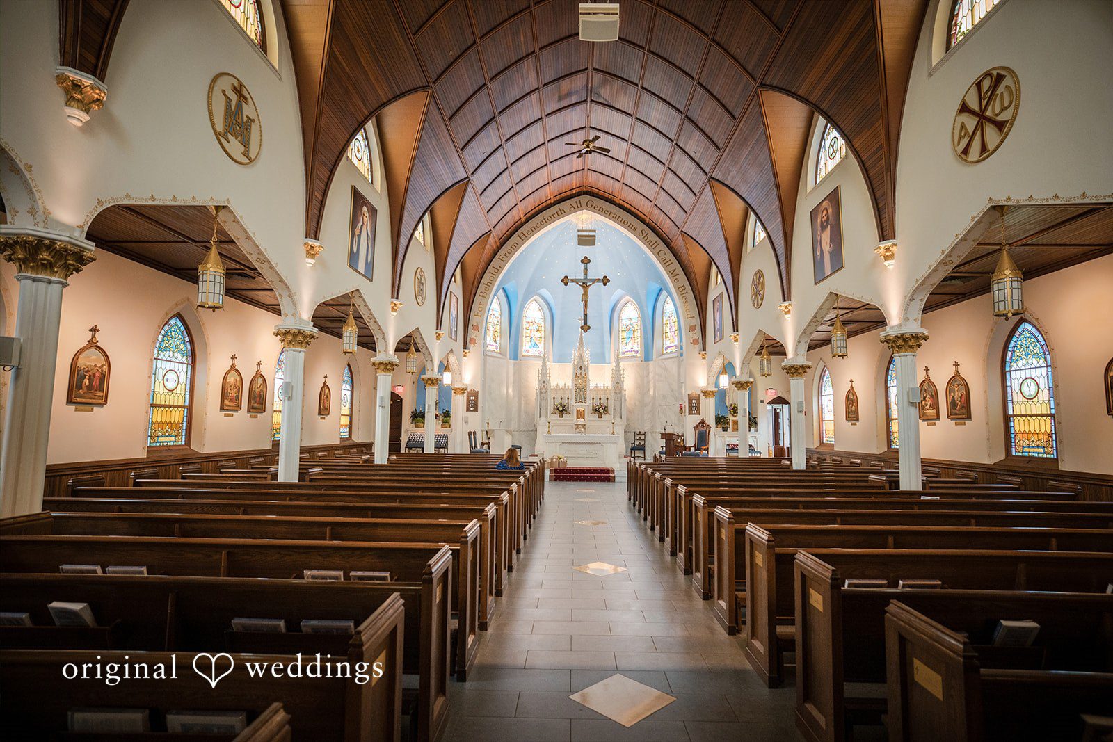 Nicole + Matt The interior aisle of a church lined with wooden pews leading toward an altar.