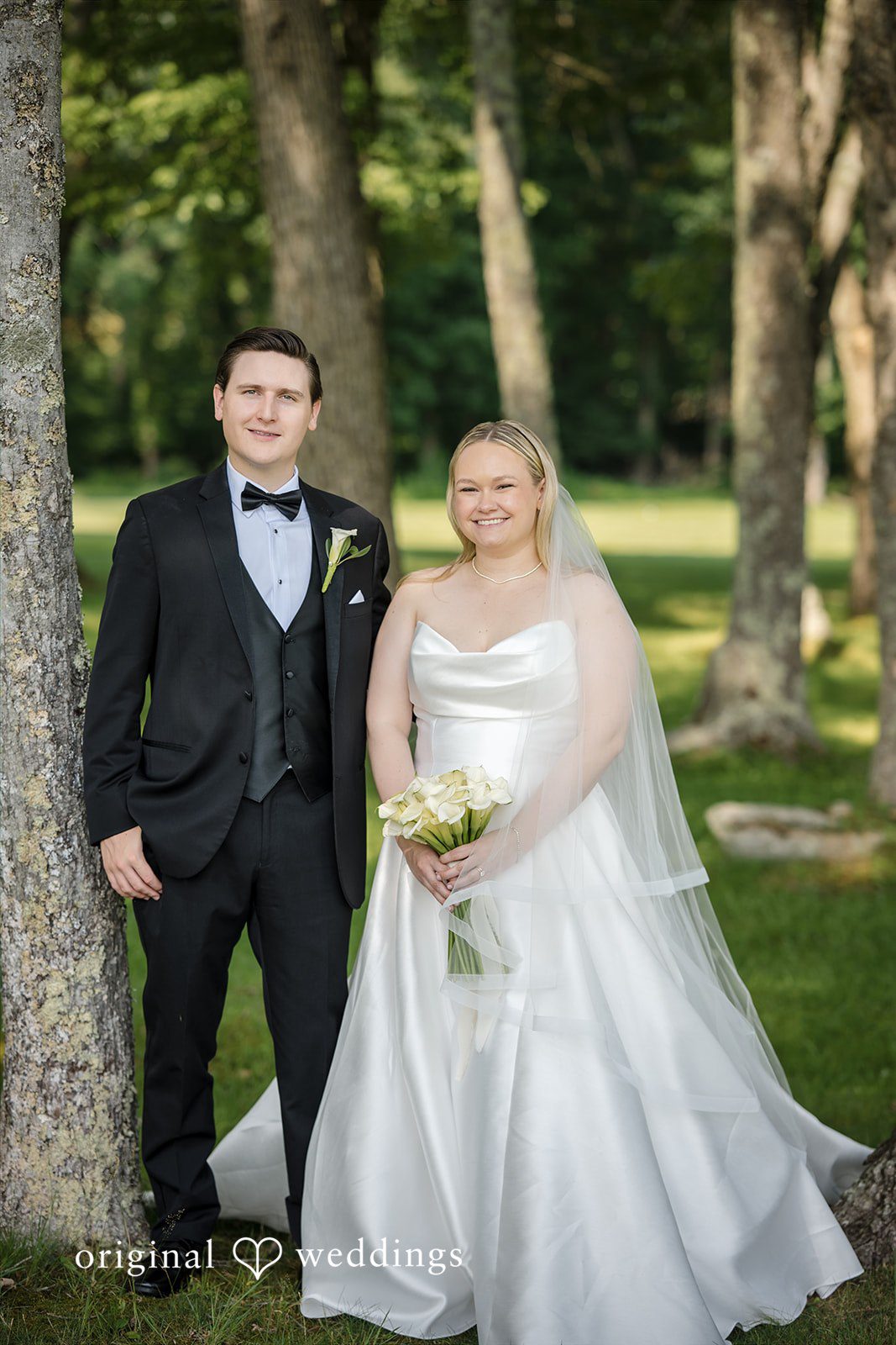 Nicole + Matt A bride and groom standing together and smiling in a grassy area with trees.