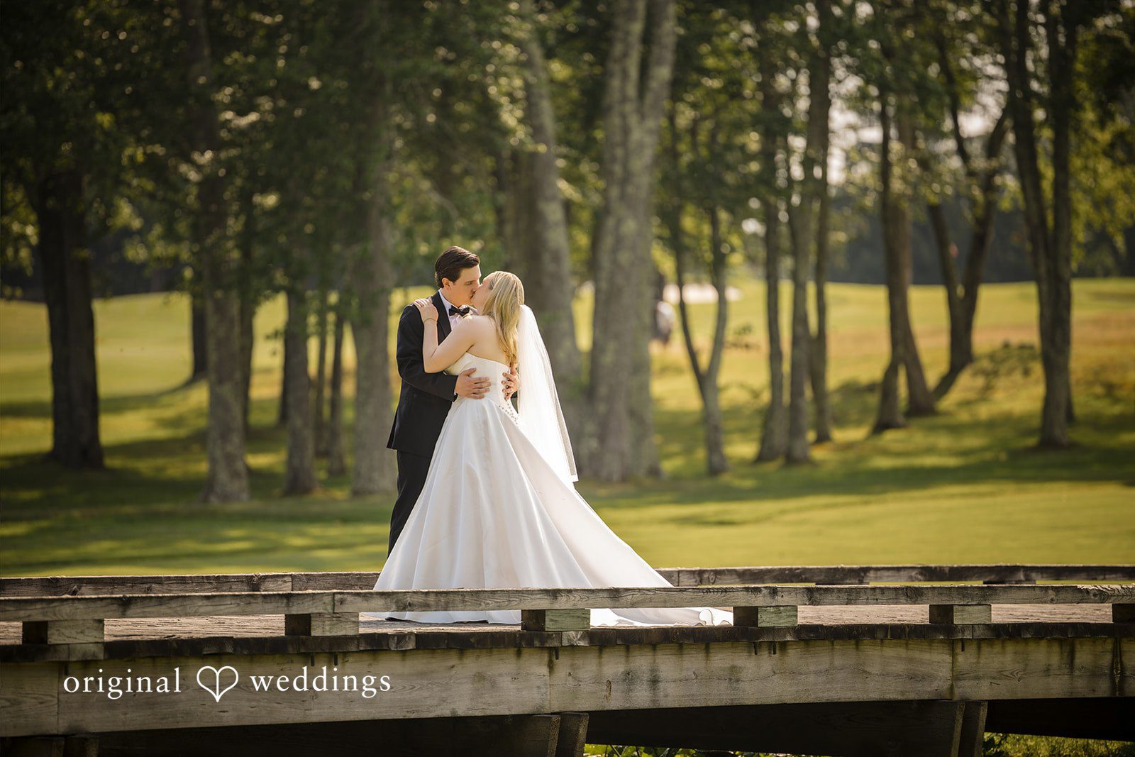 Nicole + Matt A bride and groom posing together on a wooden footbridge surrounded by trees.