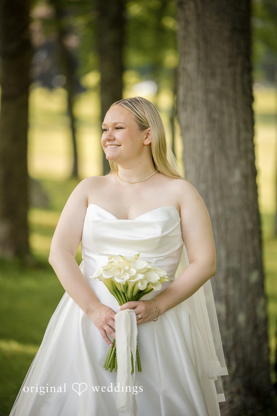 Nicole + Matt A smiling bride holding a bouquet while standing outside in front of a tree trunk.