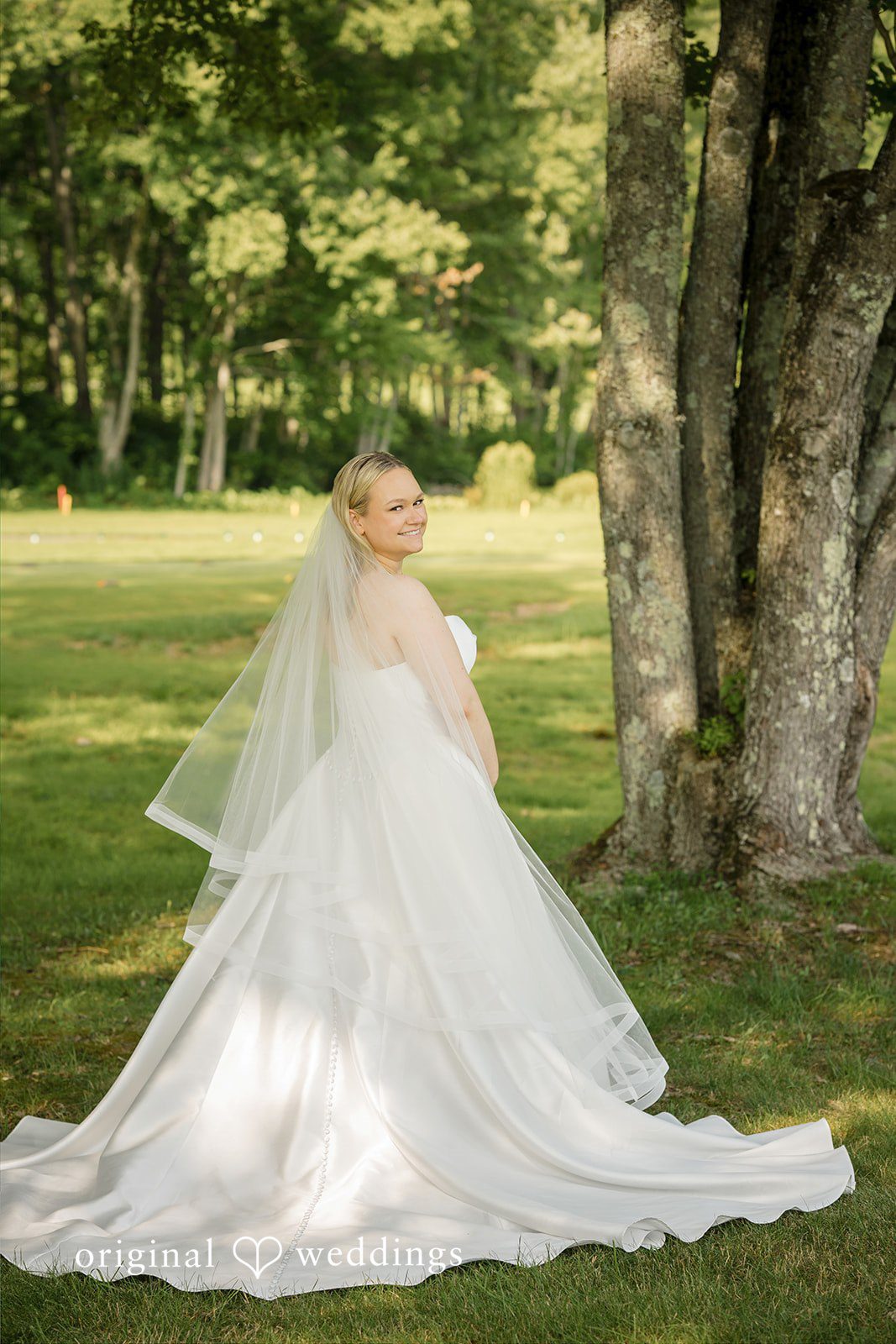 Nicole + Matt A bride in a white gown posing on a green grass lawn surrounded by trees.