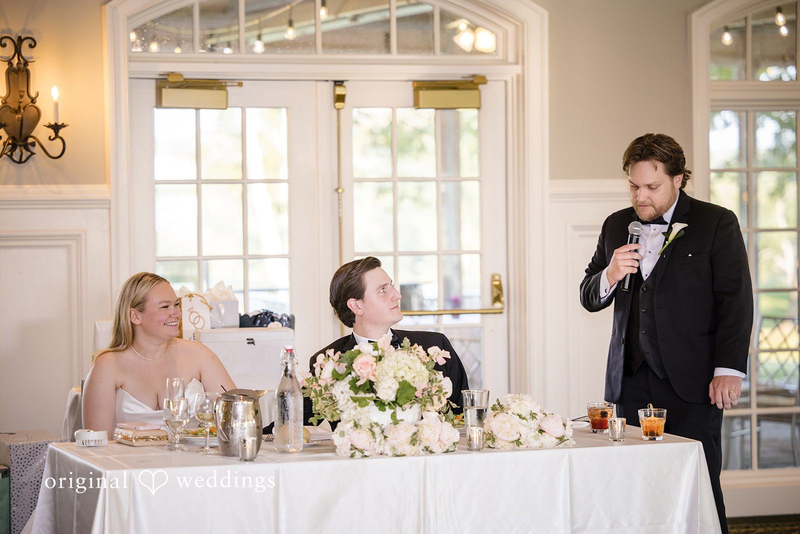 Nicole + Matt A man in a black tuxedo speaks into a microphone at a wedding reception head table while the seated bride smiles and looks on.