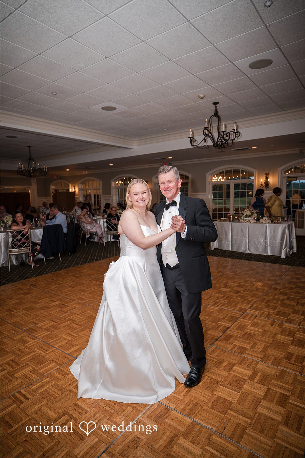 Nicole + Matt A smiling bride and a man in a black tuxedo dance together on a wooden dance floor during a wedding reception.
