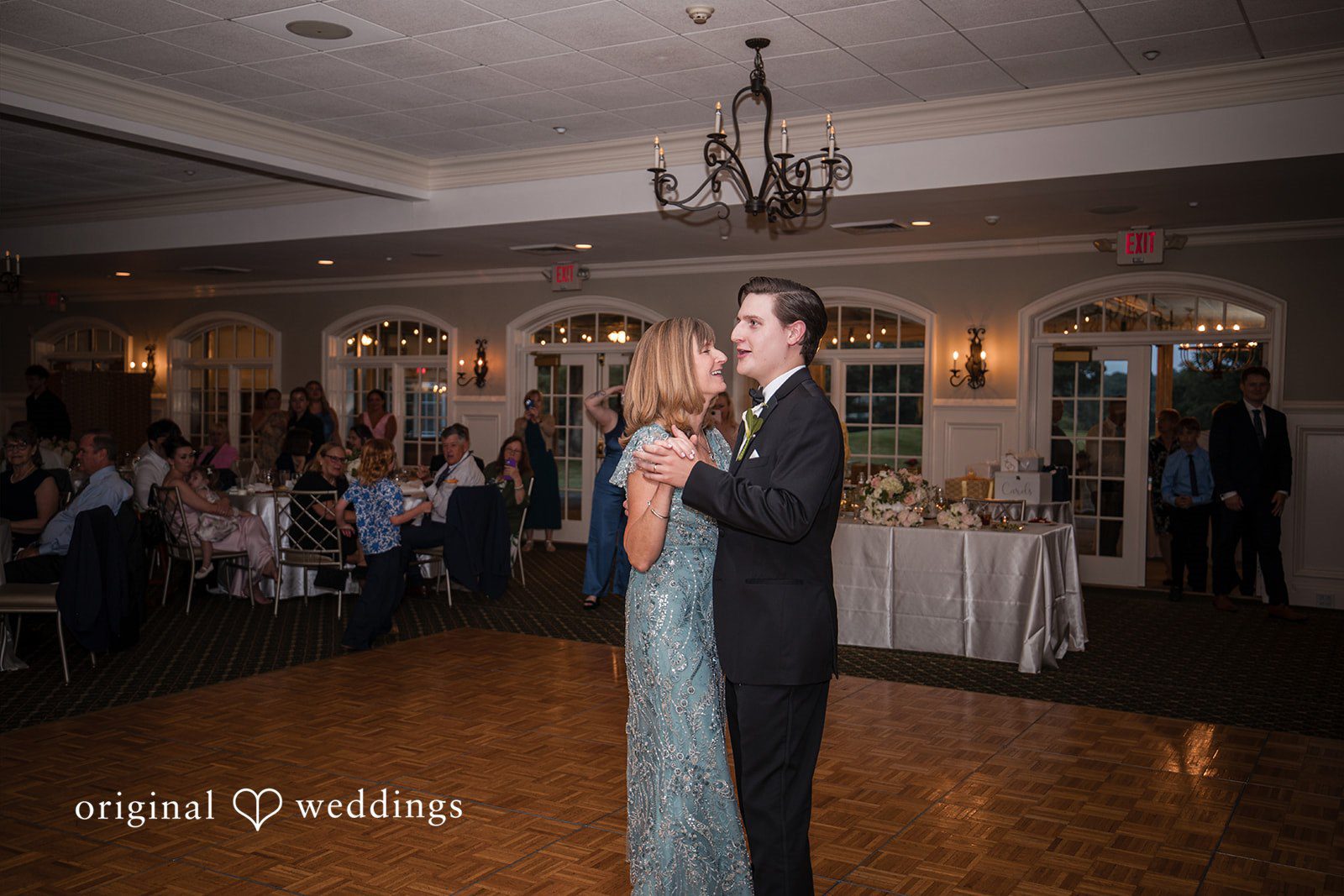 Nicole + Matt A groom in a black tuxedo dances with a woman in a sparkling blue dress on a wooden dance floor during a reception.