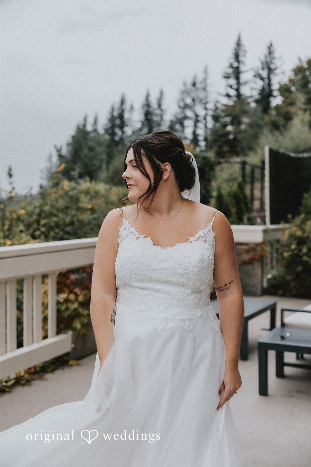 A bride posing on a balcony railing with evergreen trees in the background.