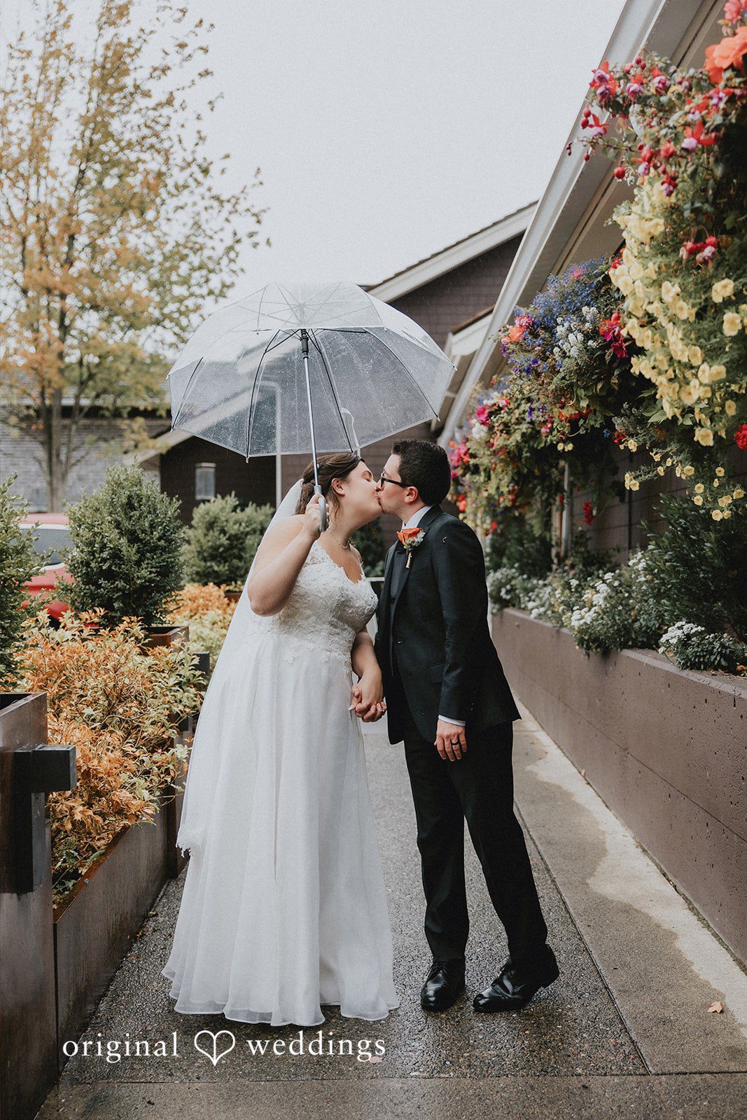 A bride and groom walking down a path under an umbrella next to a building.