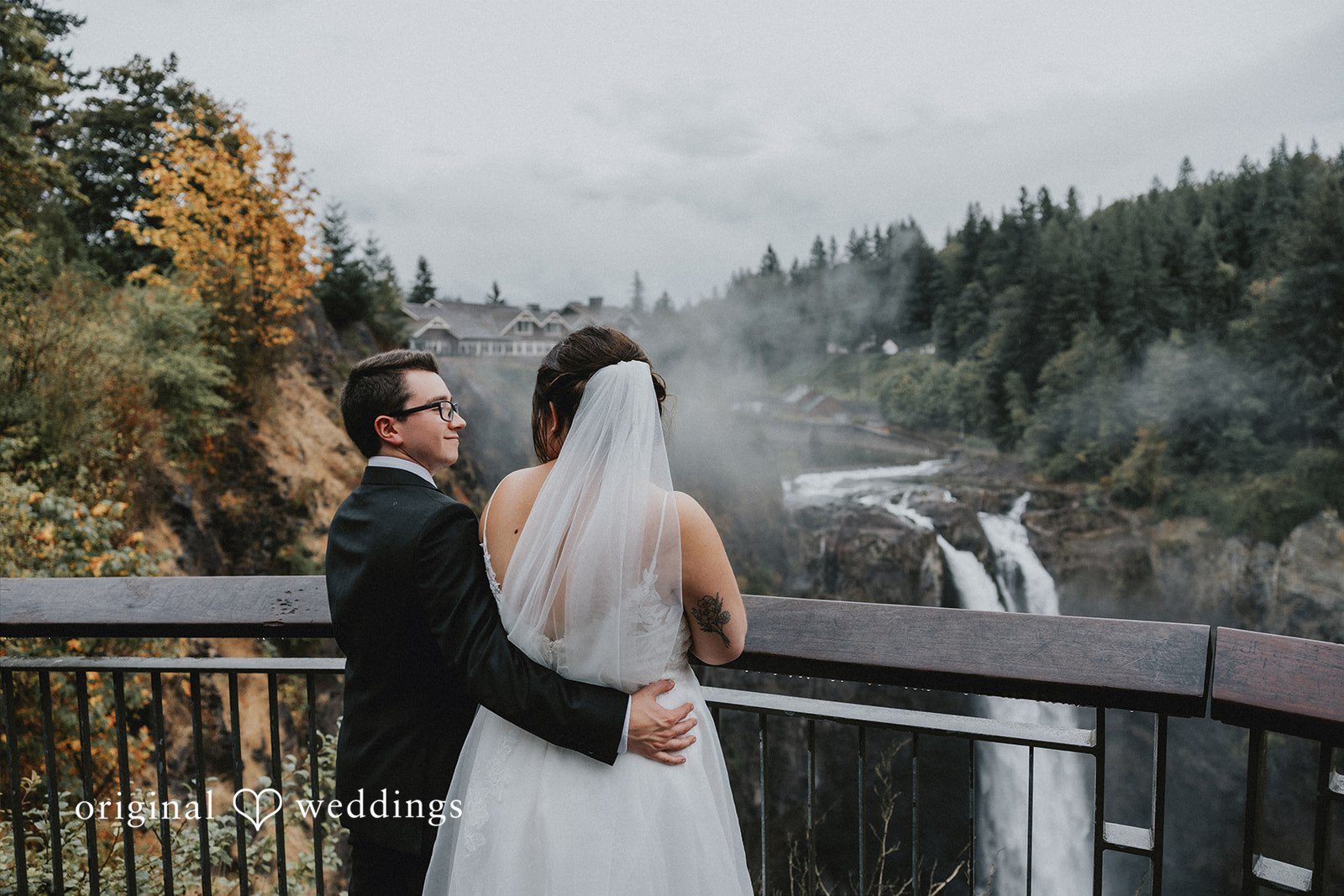 A bride and groom posing against a railing with a dense pine forest behind them.