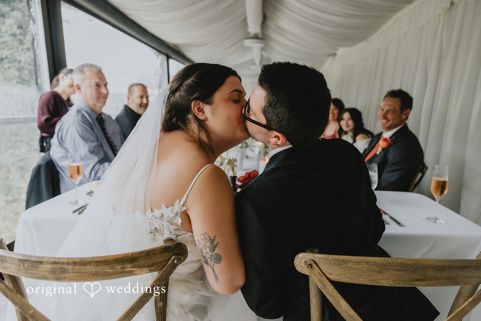 A bride and groom sitting together and kissing at a reception table.