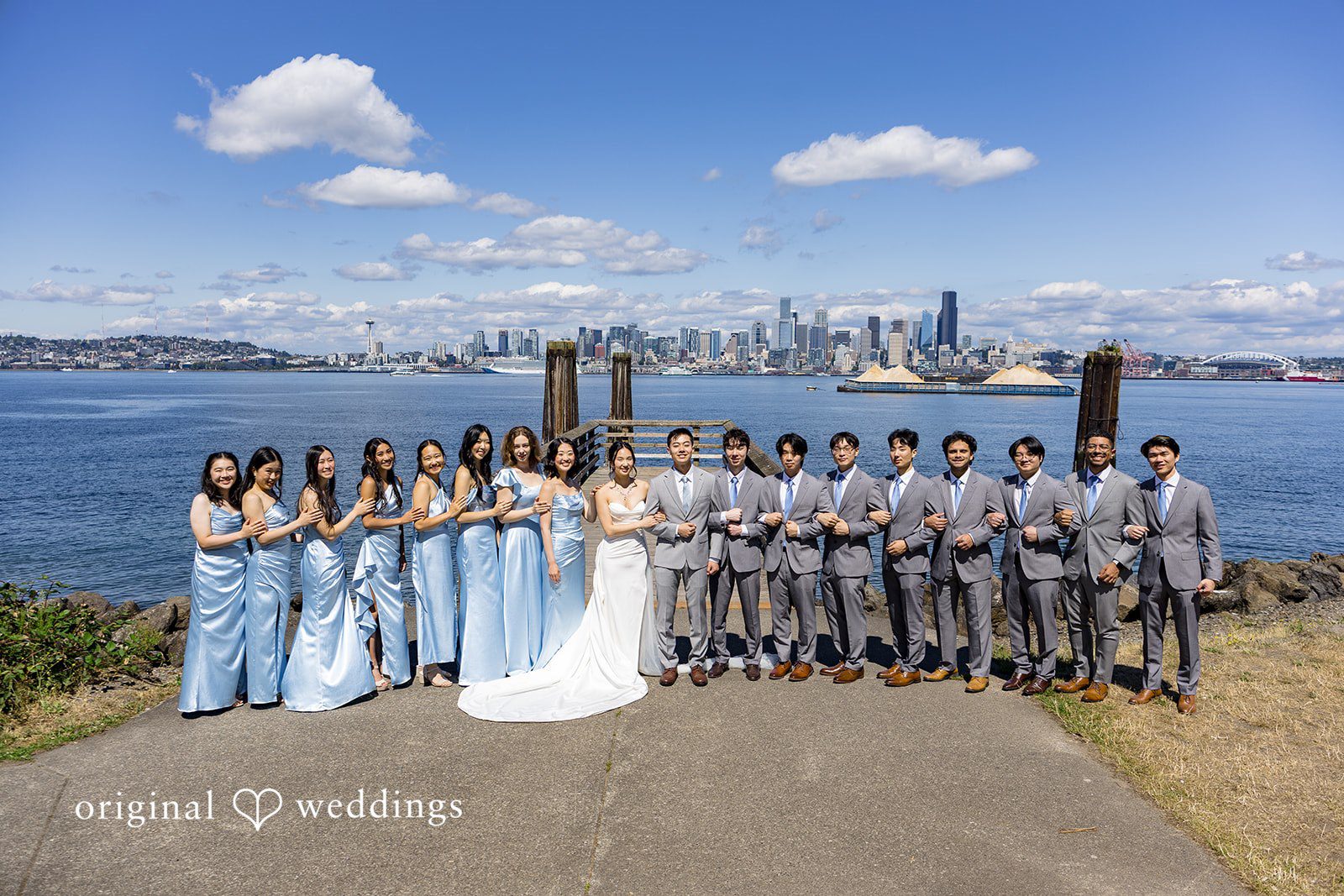 Emily + Paul A wedding party in formal attire standing in a long line posing along a scenic waterfront pier.