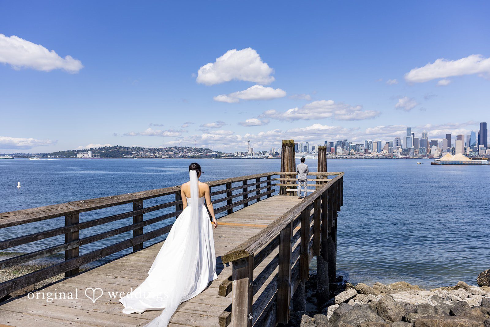 Emily + Paul A wide scenic view of a bride walking down a long wooden pier extending into the water.