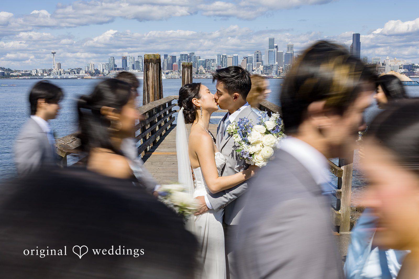 Emily + Paul A candid photo of wedding guests chatting near a wooden railing by the shore.