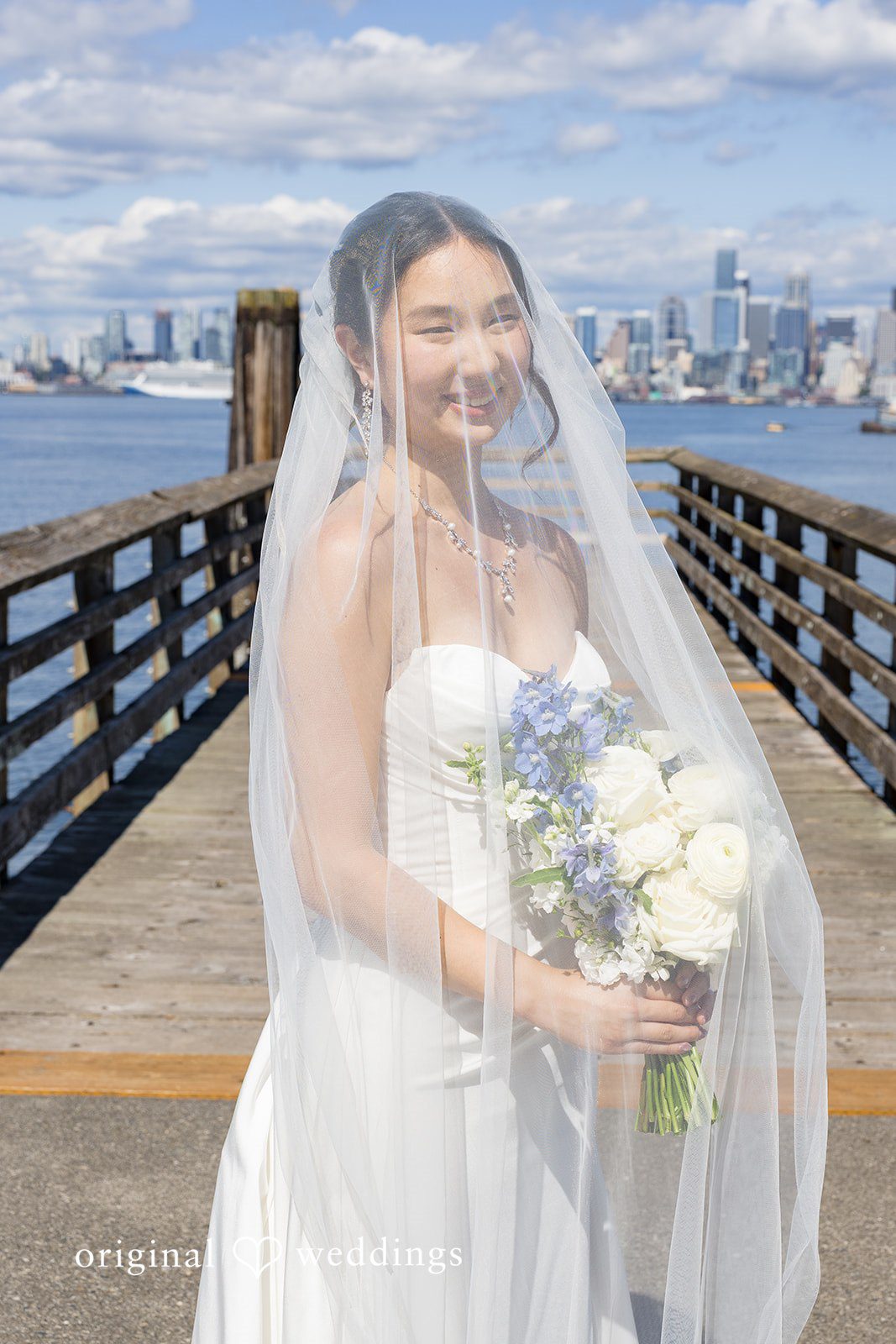 Emily + Paul A bride wearing a long sheer veil smiling as she stands against a city waterfront background.