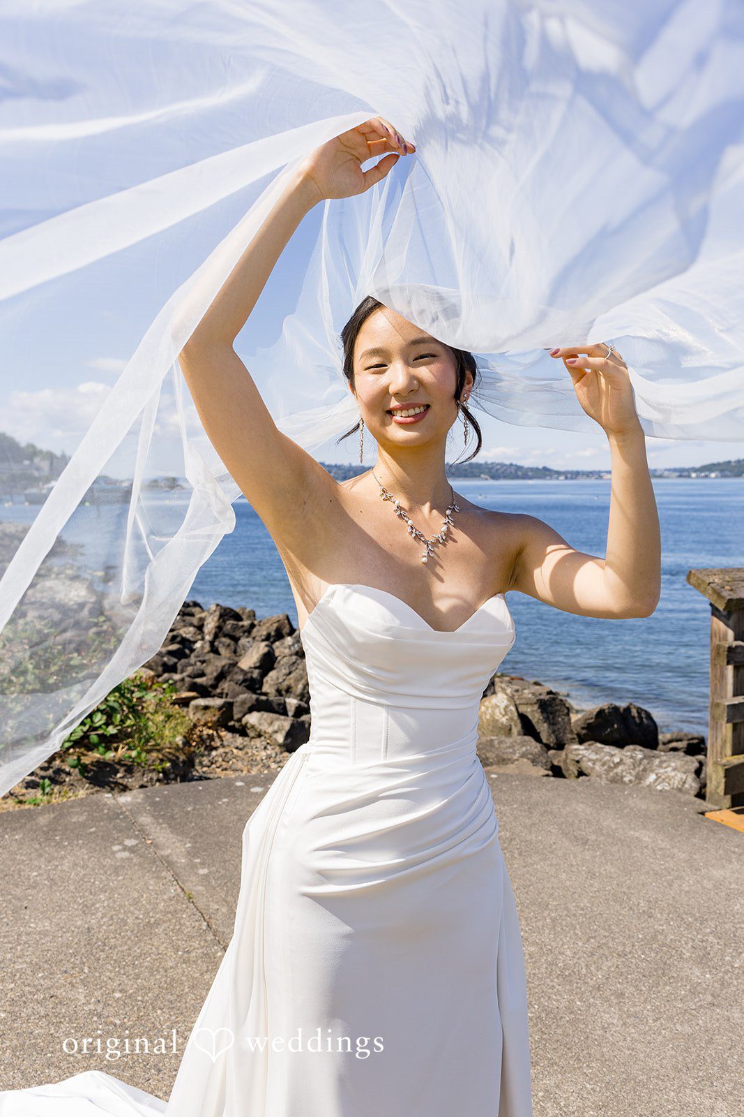 Emily + Paul A joyful bride raising her arms while holding a flowing veil in front of a rocky shoreline.