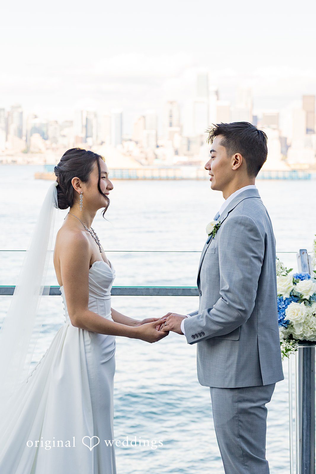 Emily + Paul A bride and groom holding hands while smiling at each other against a coastal backdrop.