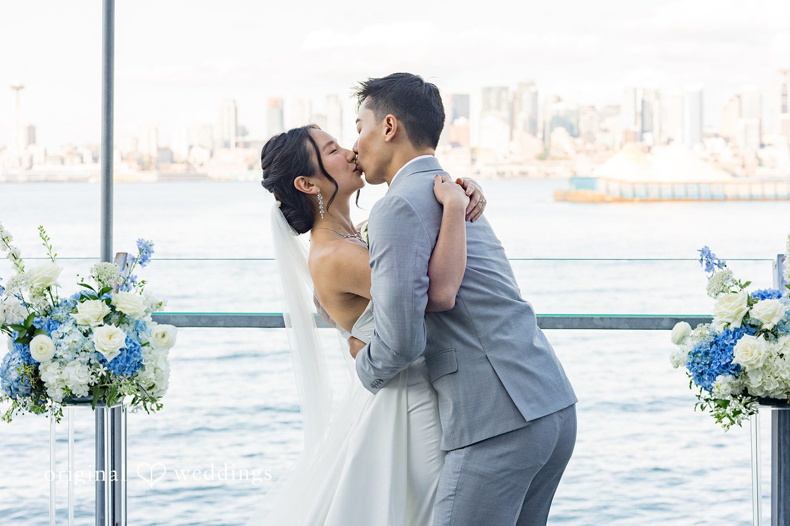 Emily + Paul A bride and groom embracing by the water with floral arrangements framing the scene.