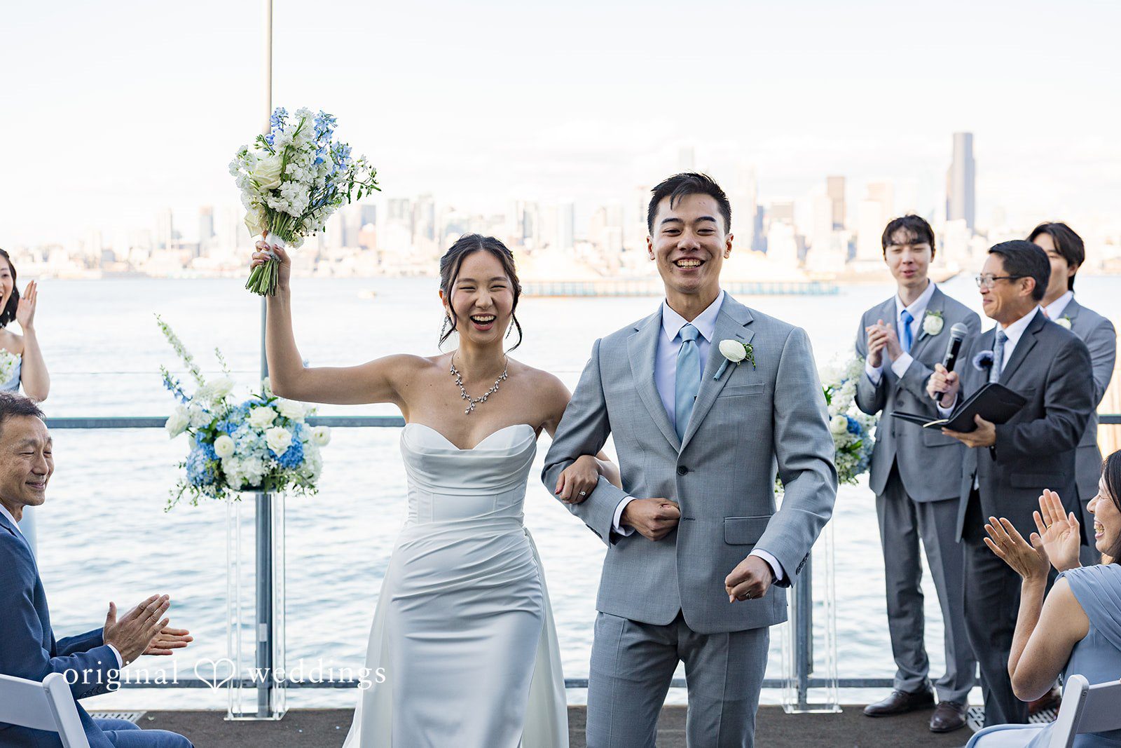 Emily + Paul A wedding party in formal wear walking and waving together along a coastal pathway.