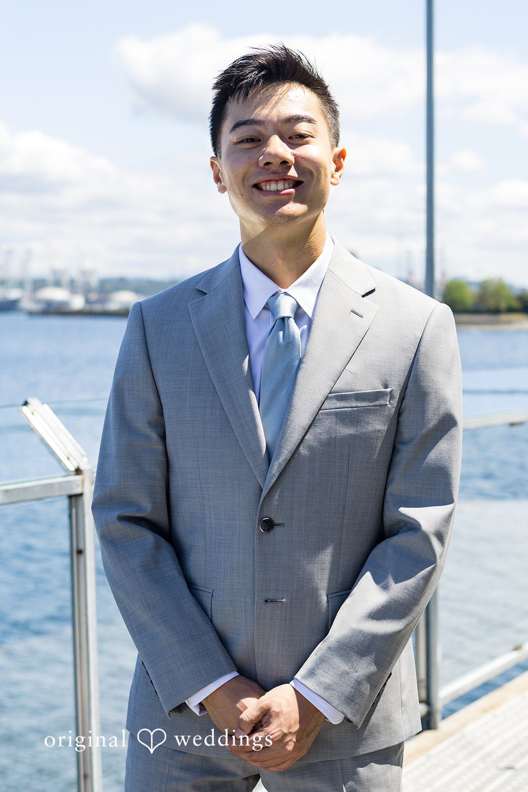 Emily + Paul A smiling groom dressed in a grey suit and blue tie posing in front of a waterfront railing.