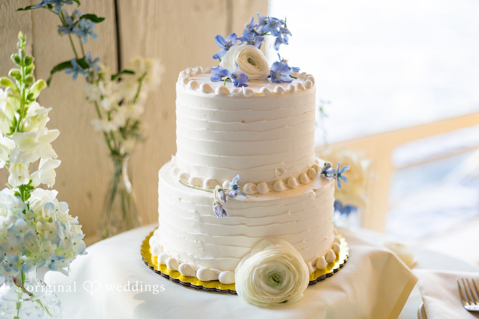 Emily + Paul A three-tiered white wedding cake decorated with gold trim and pearls sitting on a table.