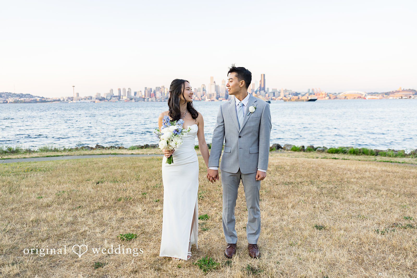Emily + Paul A bride and groom walking side by side across a green field with the ocean in the distance.