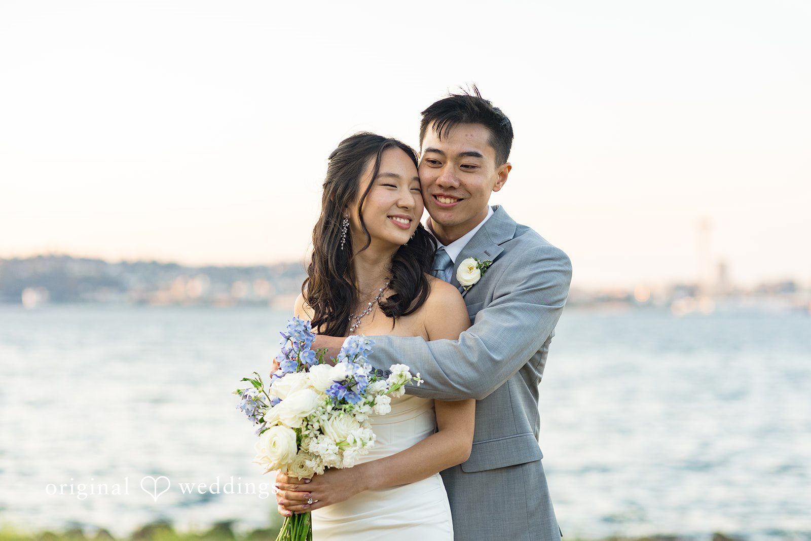 Emily + Paul A bride holding a bouquet while embracing her groom on a grassy field near the water.
