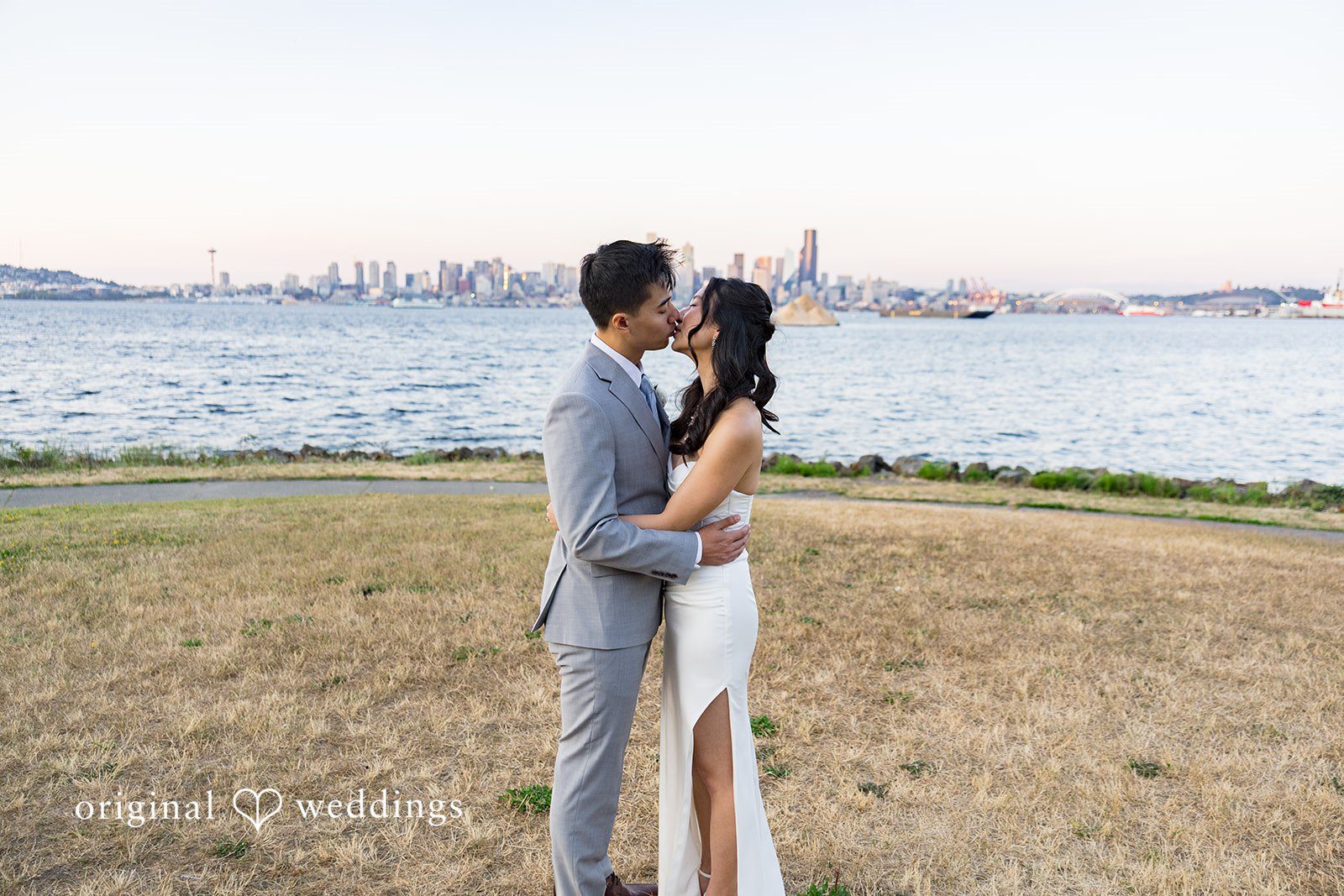 Emily + Paul A bride and groom embracing on a grassy hill next to a stone railing overlooking the ocean.