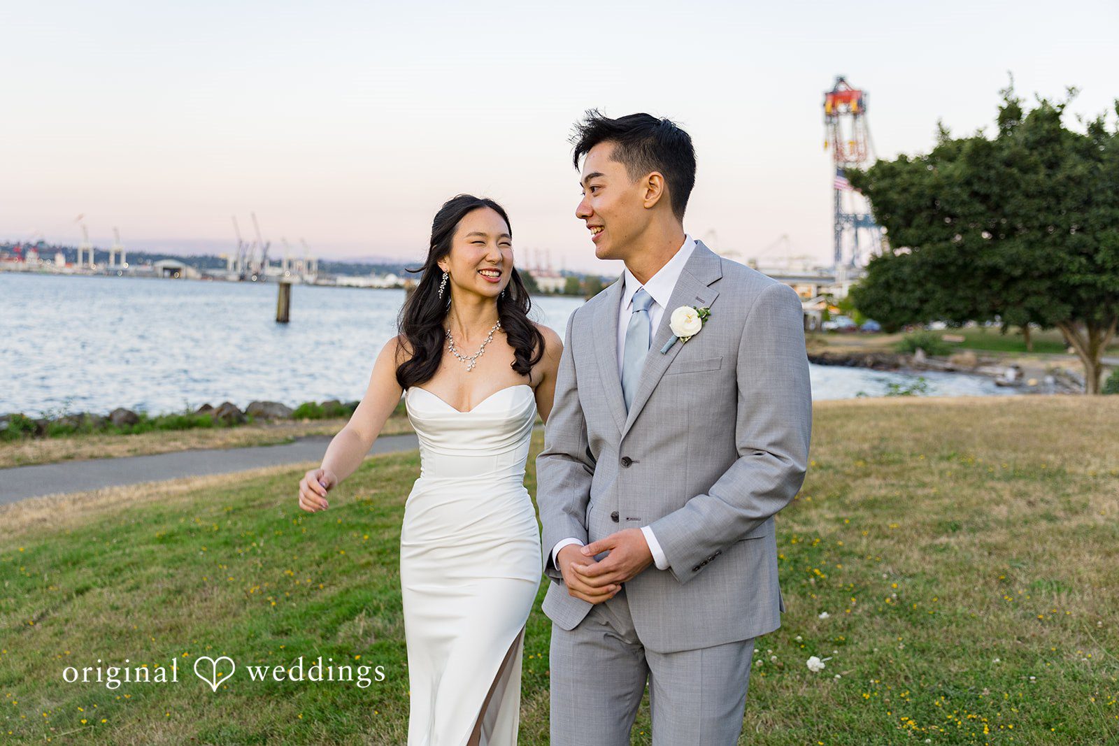Emily + Paul A bride and groom walking hand-in-hand along a grassy path near the waterfront.