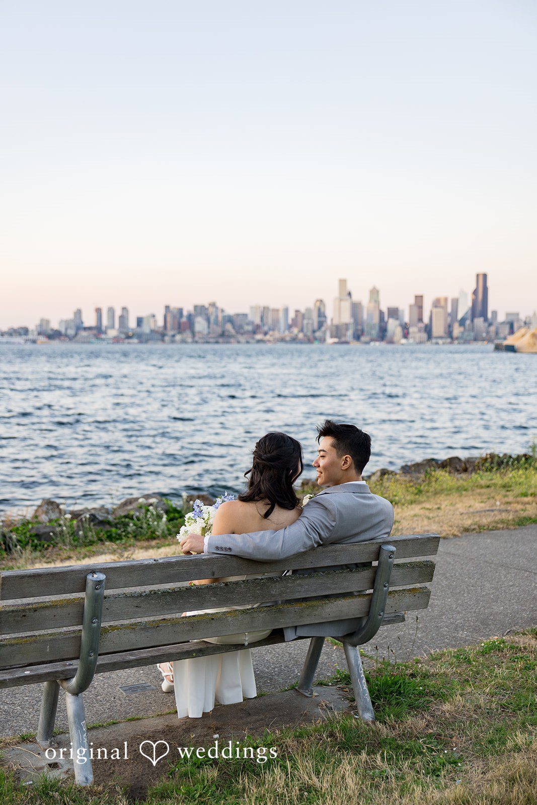 Emily + Paul A bride and groom sitting together on a wooden park bench facing a scenic waterfront view.