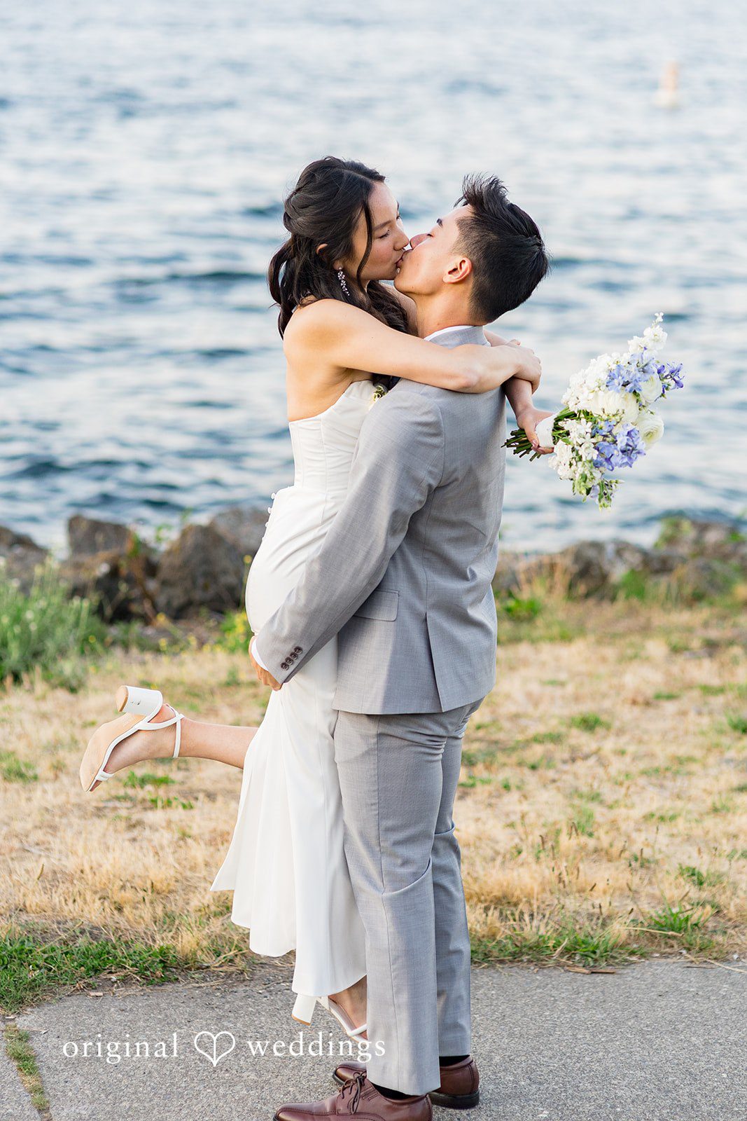 Emily + Paul A groom lifting his bride up in a joyful embrace along a rocky coastline.