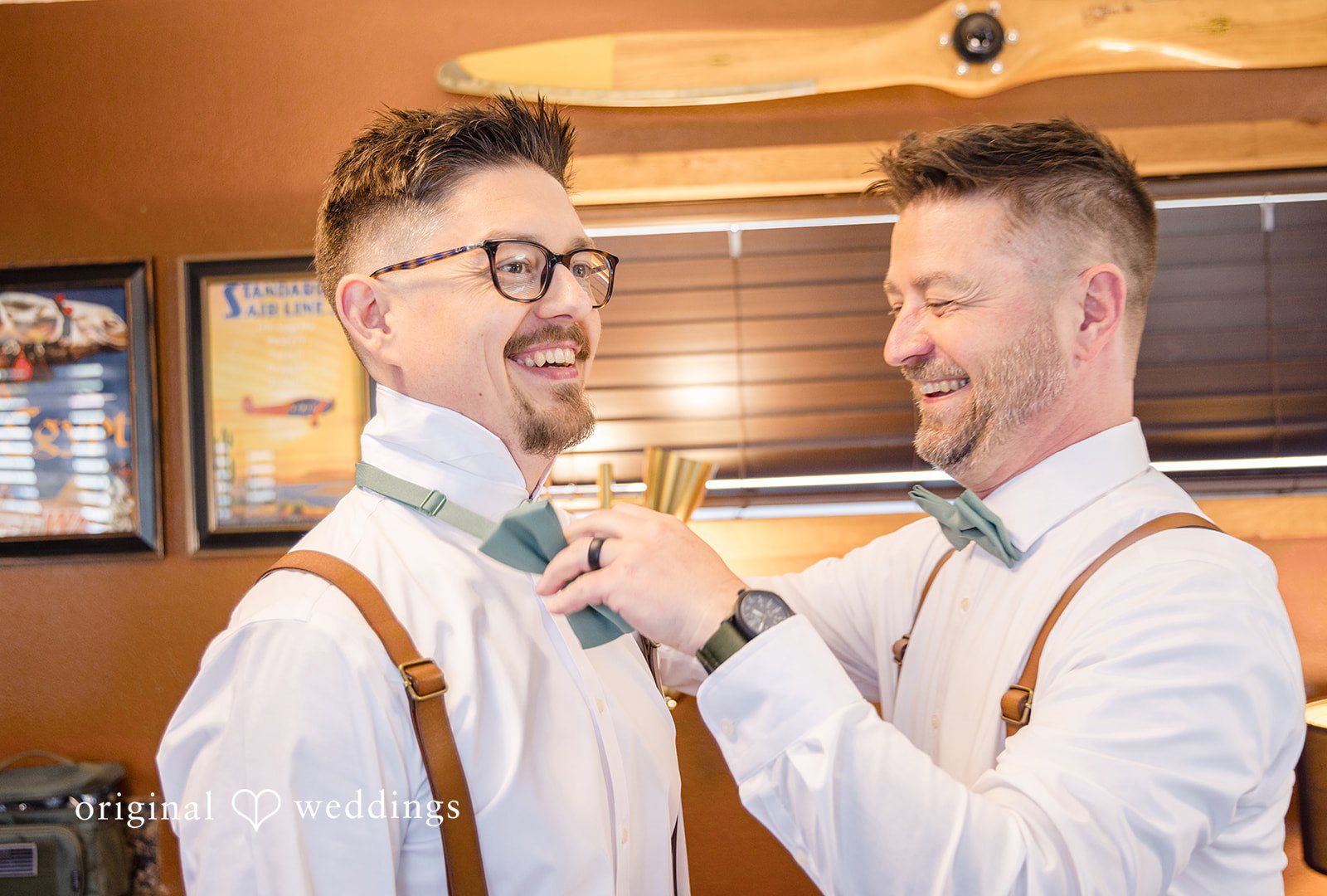 Two men in suspenders smiling as one helps the other adjust his bow tie.