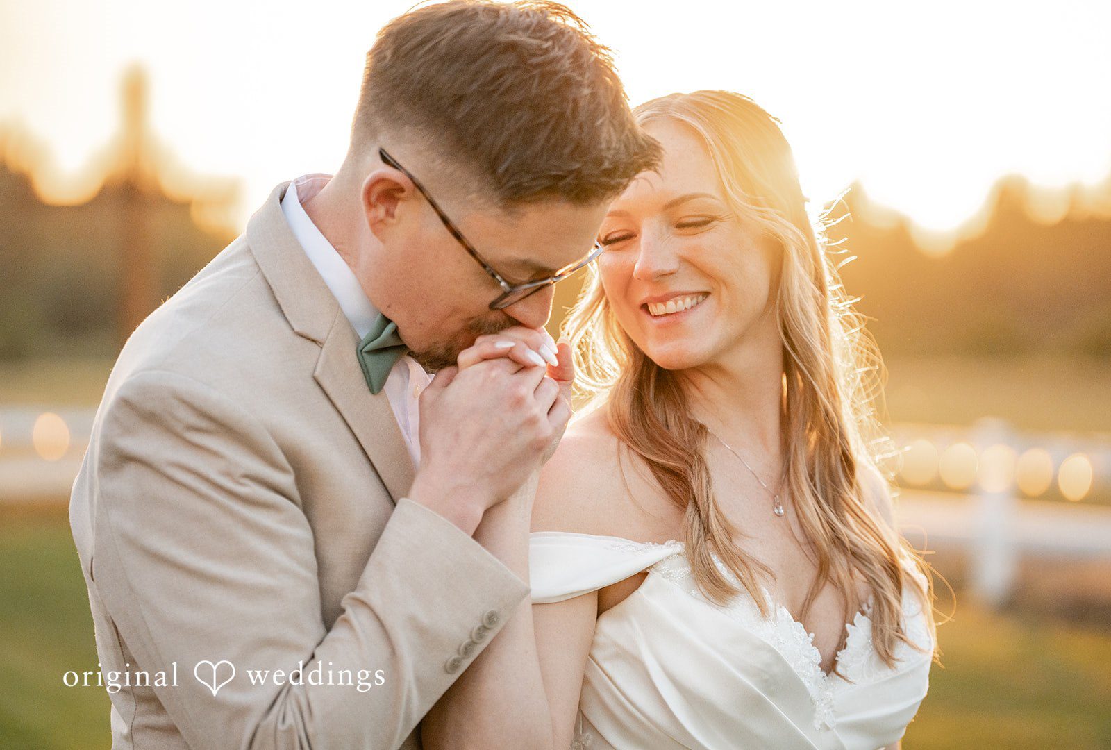 A groom tenderly kissing the hand of his smiling bride in warm sunlight.