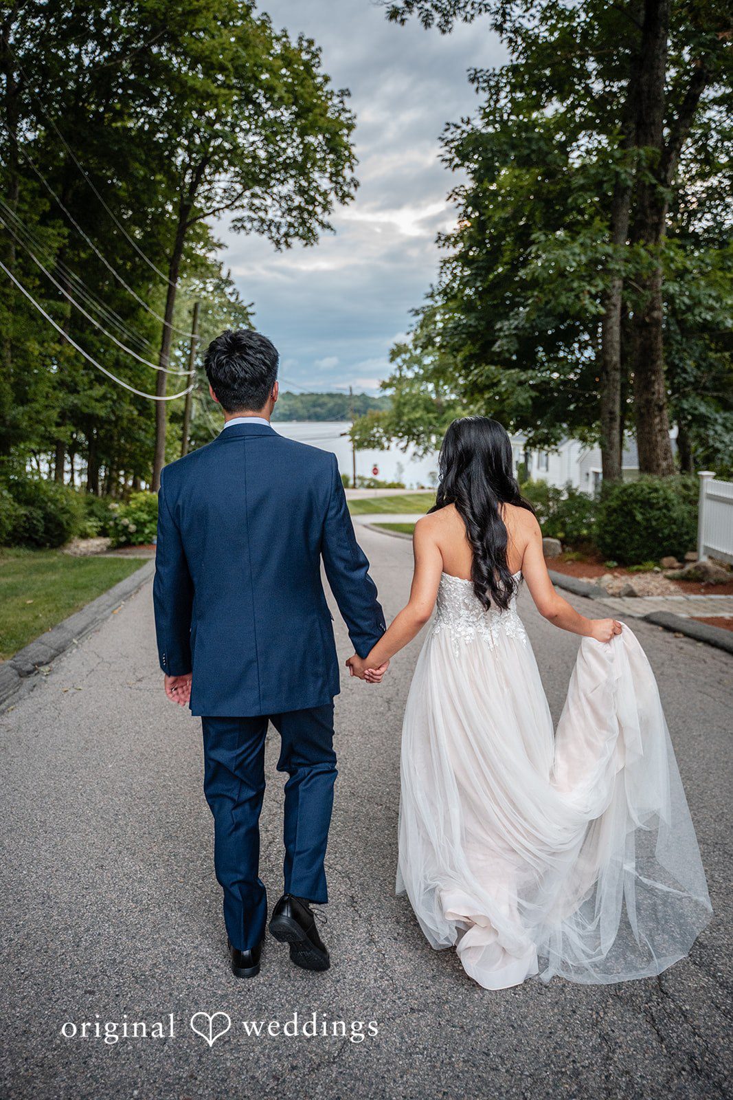 Bride and groom walking together down a paved pathway lined with trees.