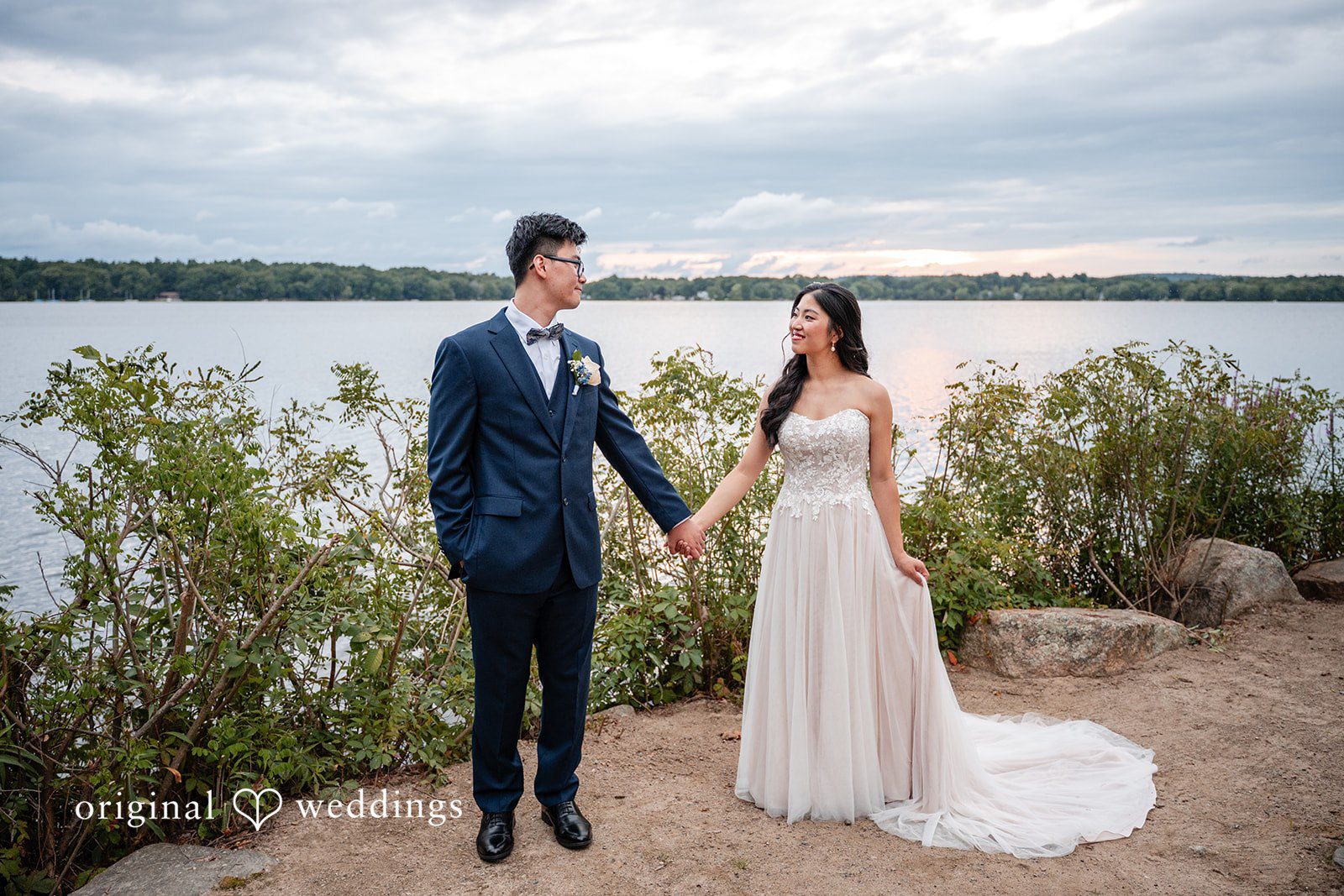 Bride and groom posing together on a rocky shoreline by the water.