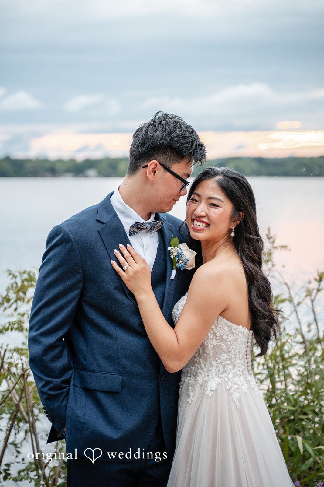 Groom in a dark suit embracing a smiling bride in front of a waterfront view.