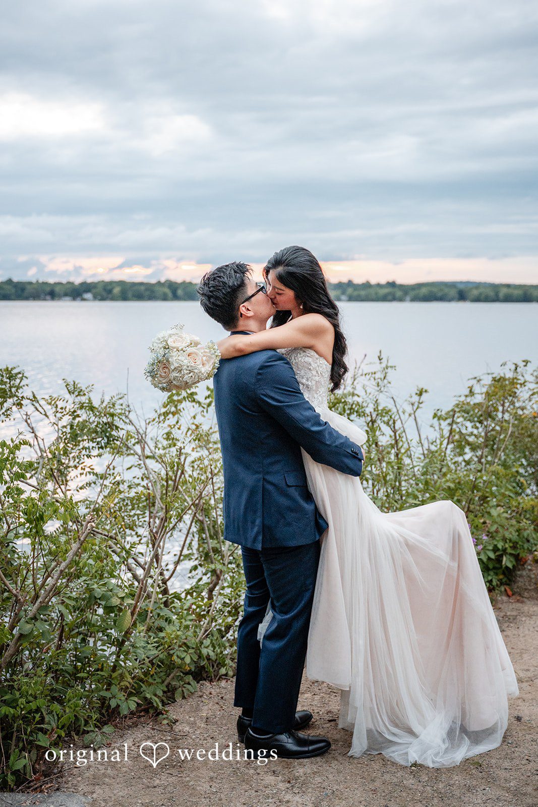 Bride and groom embracing on a grassy hill overlooking a body of water.