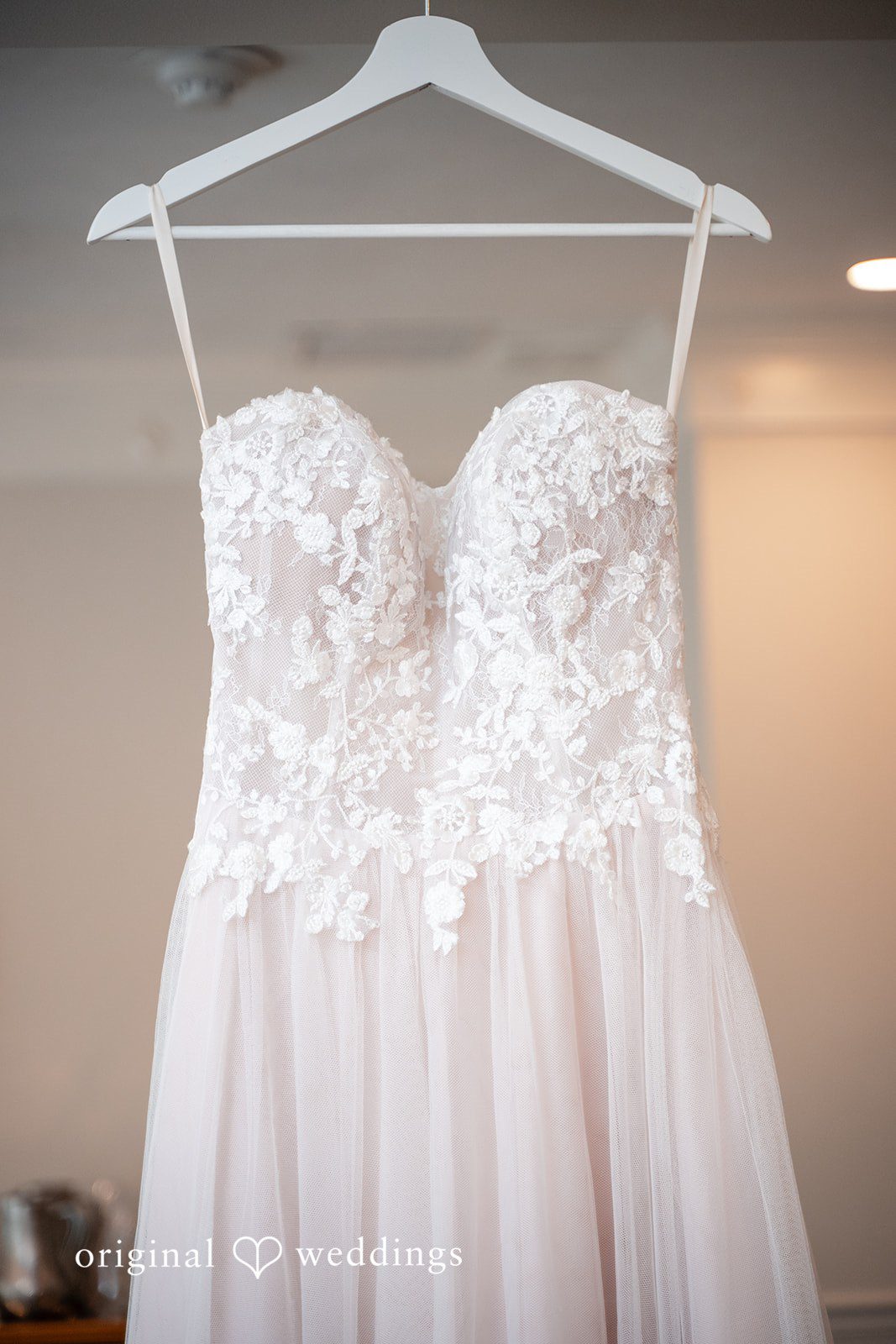 Strapless white lace wedding gown hanging on a wooden hanger.